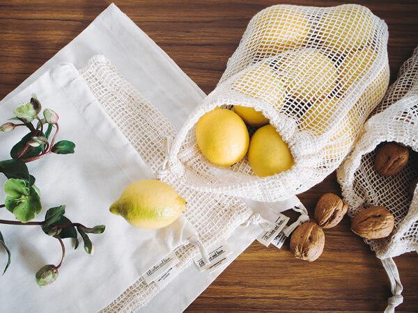 A variety of breathable mesh produce bags on a kitchen counter containing fresh fruits and vegetables like apples, oranges, carrots, and leafy greens. Each bag is tied with a drawstring, showcasing colorful produce through the mesh fabric. 