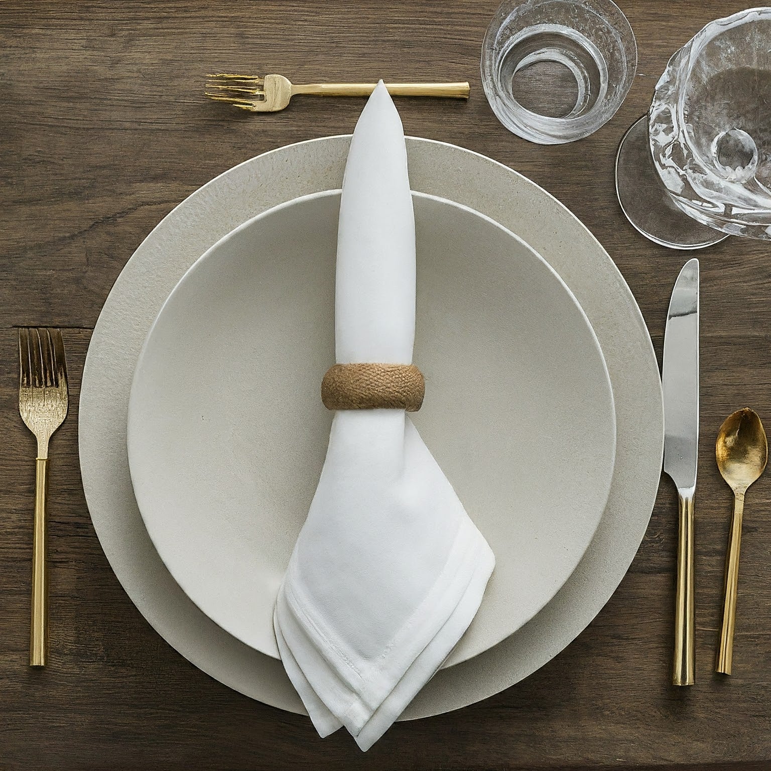 A place setting with a white napkin folded into a triangle on a white plate.  Wooden napkin rings, silver utensils, and a glass are also on the table.