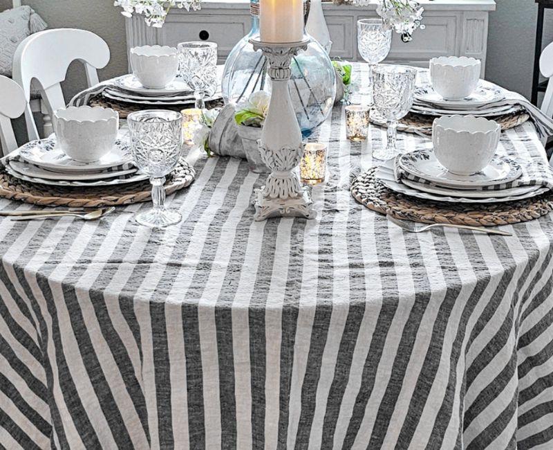 Cozy dining room with a table set for a meal, featuring a bold and colorful striped tablecloth with red, white, and blue stripes, plates, glasses, cutlery, natural light, and potted plants adding a touch of greenery