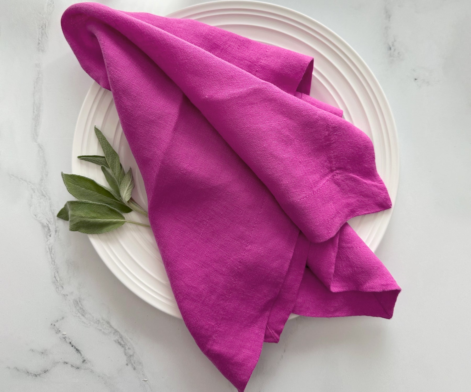 Orchid linen dinner napkin folded on a white plate with fresh sage leaves beside it.